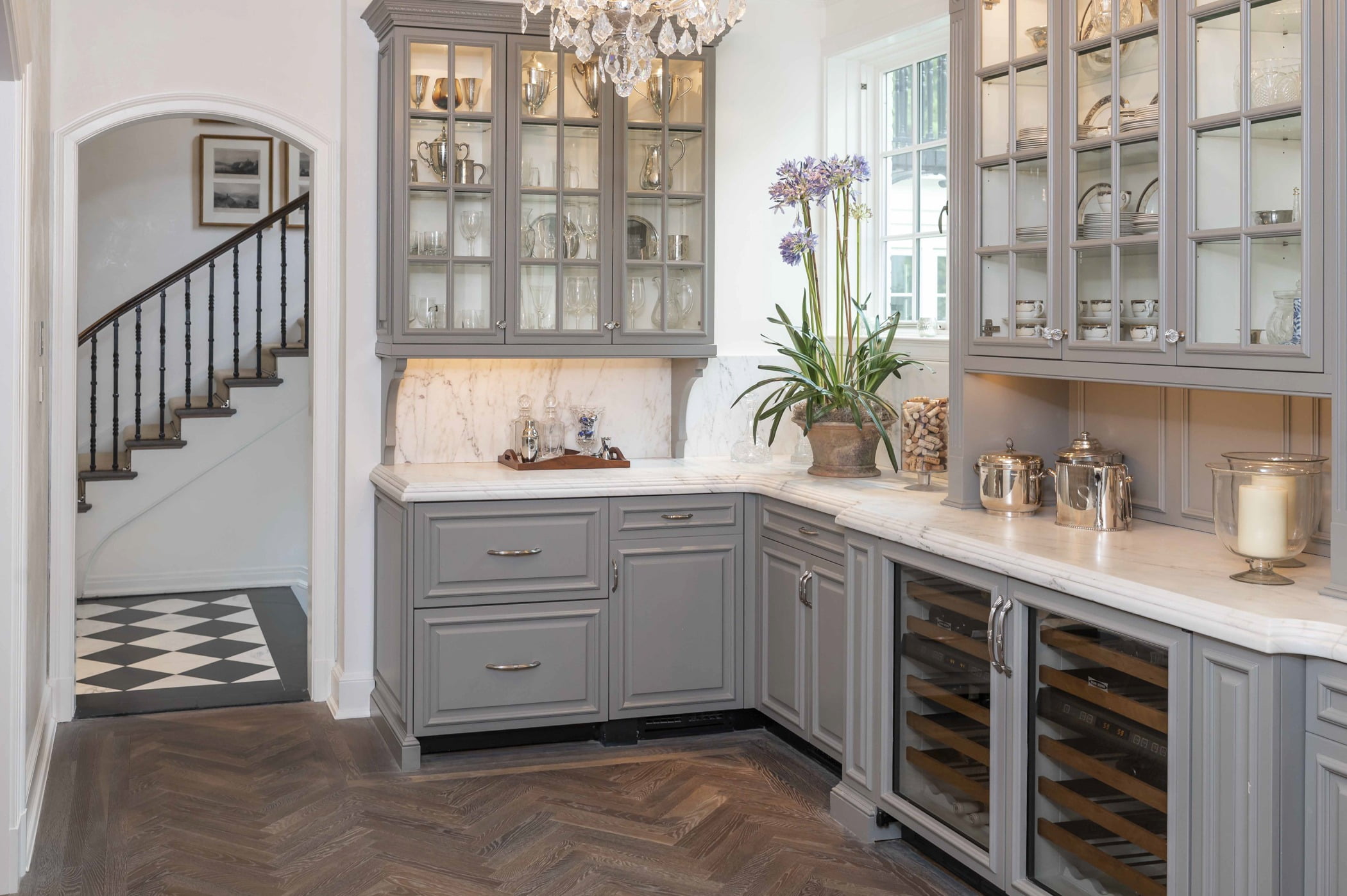 Traditional kitchen area with custom light grey cabinetry, marble countertops, ornate crystal chandelier, and dark chevron hardwood flooring (Doorway view)